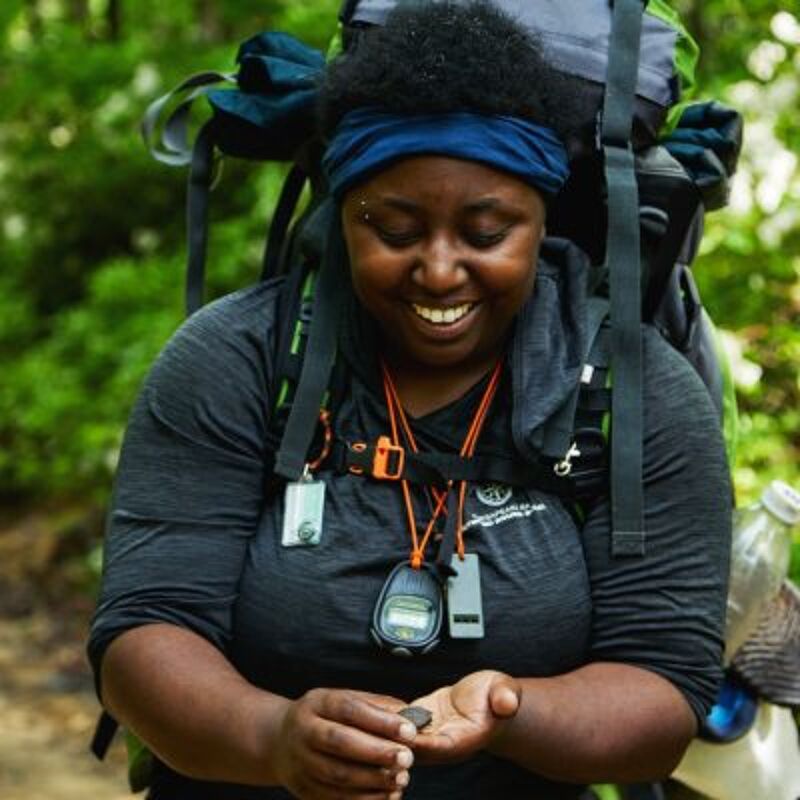 A woman with a backpack is looking down at something in her hands. She is wearing a blue headband and has a piercing above her left eyebrow. She is wearing a gray long-sleeved shirt and an orange lanyard. She is smiling. The background is blurry, but it appears to be a forest.
